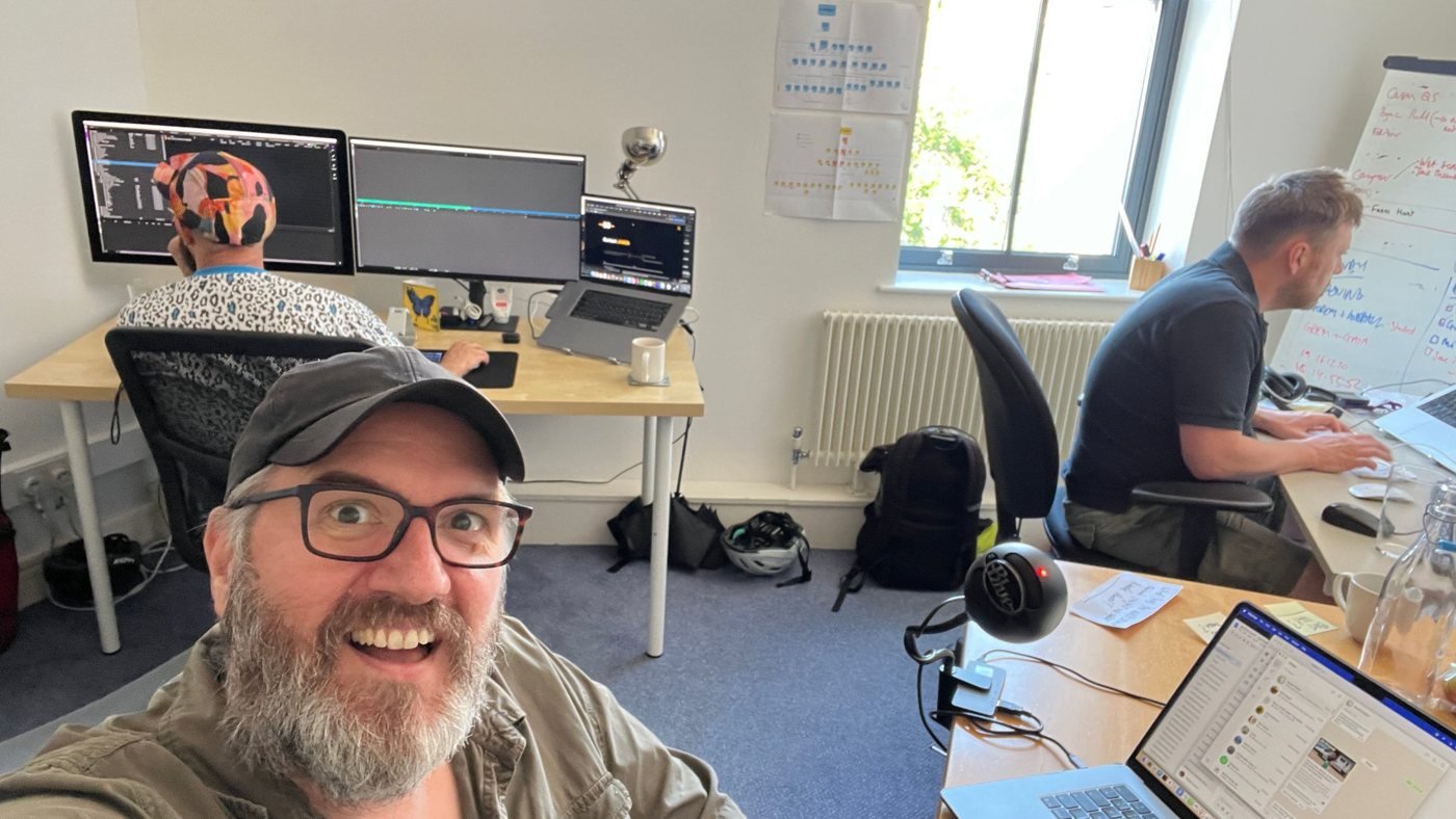 Andy grinning at the camera in a busy edit office, two colleagues working at their screens behind him